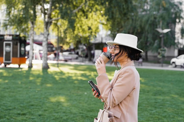 Mulher de negócios está bebendo café ao ar livre no parque. Garota usava chapéu branco, vestido e jaqueta.