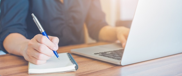 Mulher de negócios está escrevendo em um caderno com uma caneta e usando um laptop para trabalhar no escritório.