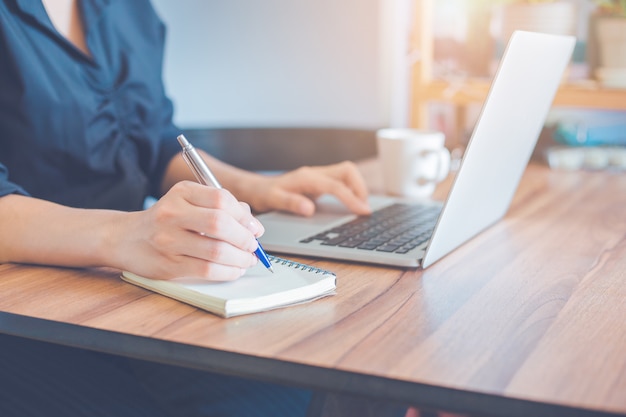 Mulher de negócios está escrevendo em um caderno com uma caneta e usando um laptop para trabalhar no escritório.