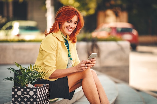 Mulher de negócios está sentada nas escadas e mensagens de texto no smartphone ao lado de uma caixa cheia de seus pertences pessoais do escritório logo depois que ela desiste do emprego.