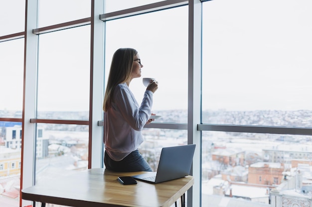 Mulher de negócios europeia usando um computador portátil enquanto trabalhava O conceito de uma mulher moderna e bem sucedida Jovem linda mulher sorridente com café sentado em um café Interior do escritório aberto