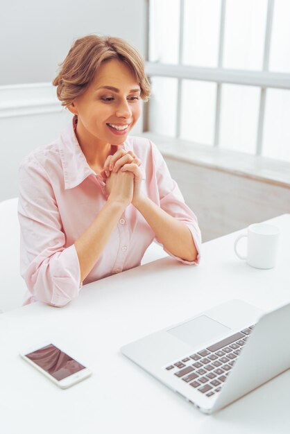 Mulher de negócios feliz atraente na camisa clássica está usando um laptop enquanto trabalha no escritório