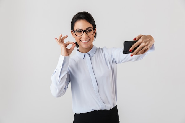 Mulher de negócios feliz e animada elegantemente vestida de pé isolada sobre uma parede branca, tirando uma selfie