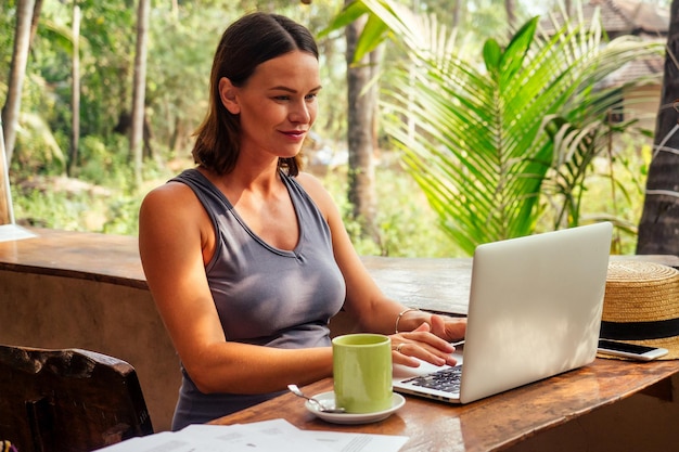 Mulher de negócios feliz e bem-sucedida usando laptop e telefone como freelancer na praia paradisíaca tropical Garota xícara chá café trabalho freelancer sentado em um café de verão trabalho remoto oceano índico