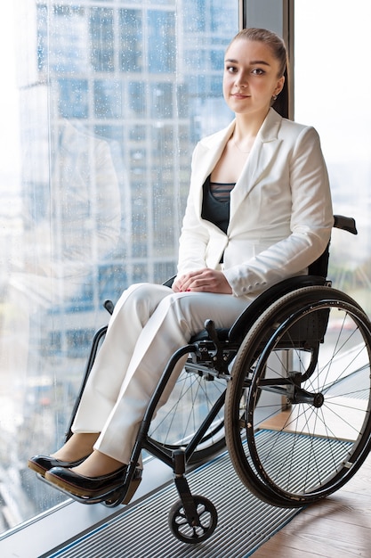 Mulher de negócios feliz e confiante em uma cadeira de rodas contra a superfície de uma janela panorâmica com vista para os arranha-céus e uma grande cidade