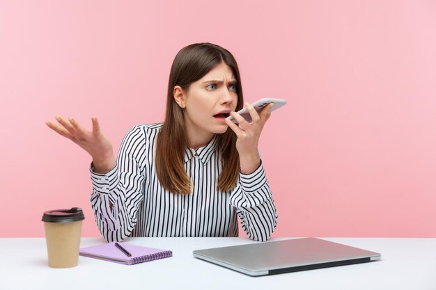 Mulher de negócios frustrada irritada na camisa listrada deixando mensagem de voz segurando o telefone perto da boca sentado no local de trabalho dando comando ao assistente de voz Tiro de estúdio interior isolado no fundo rosa