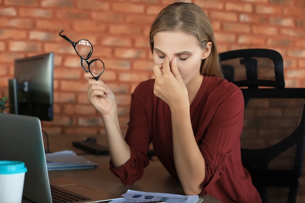 Mulher de negócios frustrada parecendo exausta enquanto está sentada em seu local de trabalho e carregando os óculos na mão.