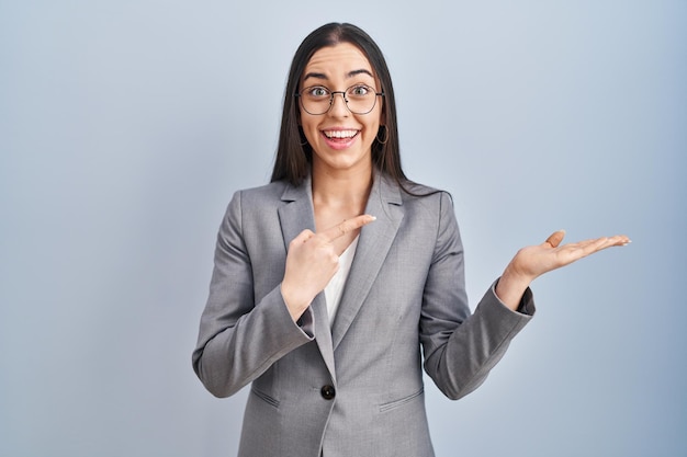 Mulher de negócios hispânica usando óculos espantada e sorrindo para a câmera enquanto apresenta com a mão e aponta com o dedo.