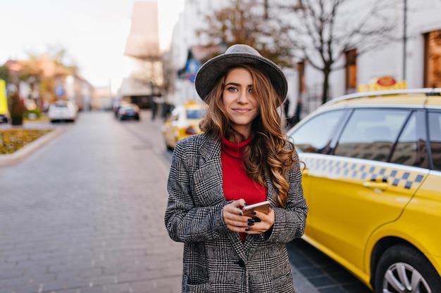Mulher de negócios incrível com casaco de tweed andando pela estrada perto de táxi amarelo. Ainda bem que mulher de cabelos compridos com chapéu, posando na rua, segurando o smartphone e olhando com interesse.
