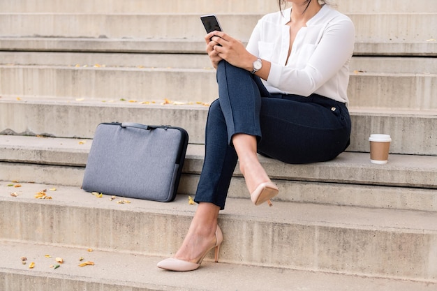 Mulher de negócios irreconhecível consultando seu telefone sentada com as pernas cruzadas em escadas de concreto, conceito de empreendedor digital e estilo de vida urbano