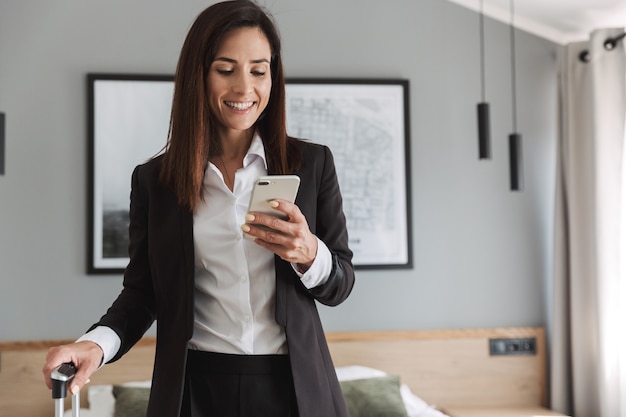 mulher de negócios jovem alegre bonita em roupas formais dentro de casa em casa com mala usando telefone celular.