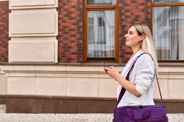 Mulher de negócios jovem andando com celular ao longo da rua da cidade, vista de perfil, espaço de cópia de fundo de construção de tijolos