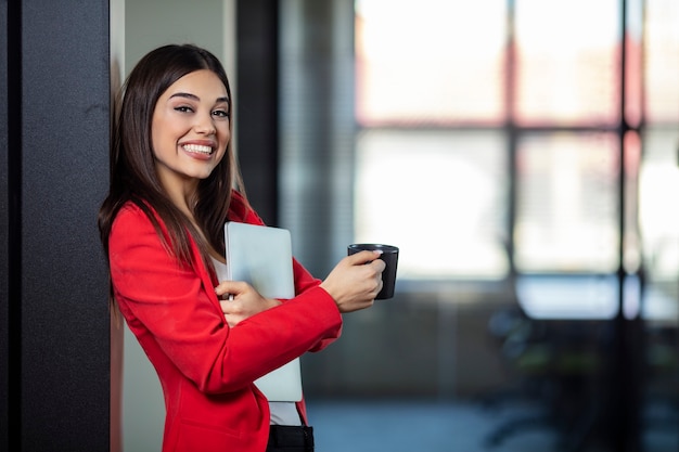 Mulher de negócios jovem bonita sorrindo e segurando o laptop e a xícara de café.