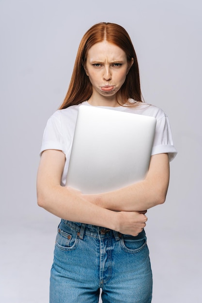 Mulher de negócios jovem chateada frustrada ou estudante segurando o computador portátil e olhando para a câmera no fundo cinza isolado. Modelo de senhora bonita com cabelo vermelho emocionalmente mostrando expressões faciais.