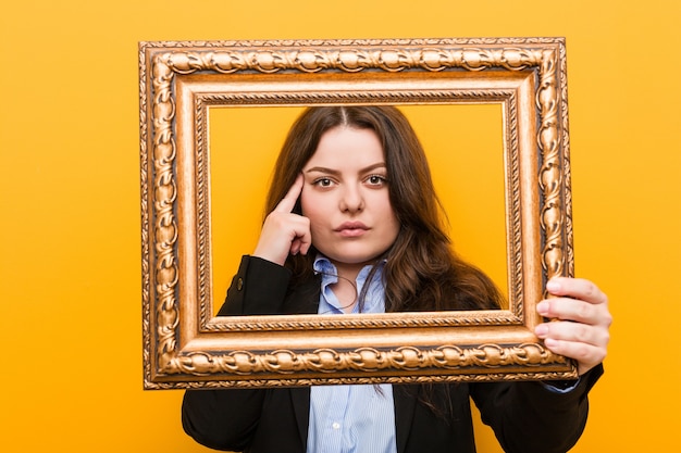 Foto mulher de negócios jovem curvilínea plus size segurando uma moldura apontando sua têmpora com o dedo, pensando, focada em uma tarefa.