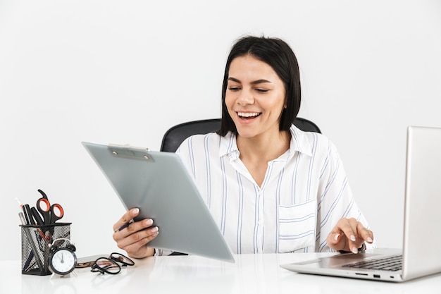 Mulher de negócios jovem e atraente sentada na mesa isolada na parede branca, trabalhando com um computador laptop e documentos