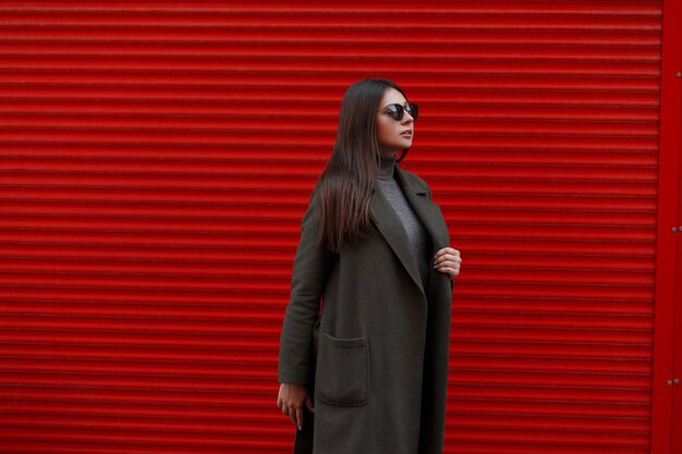 Mulher de negócios jovem e bonita elegante com óculos de sol da moda e um casaco da moda perto do portão de metal vermelho