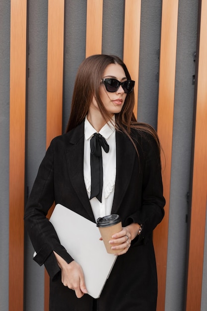 Foto mulher de negócios jovem e bonita elegante com óculos de sol em roupas de negócios elegantes com camisa e gravata com um laptop e uma xícara de café em pé perto de uma parede moderna dourada gerente de rh