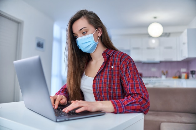 Mulher de negócios jovem em uma máscara de proteção médica trabalha em casa, no computador, durante o auto-isolamento e quarentena.