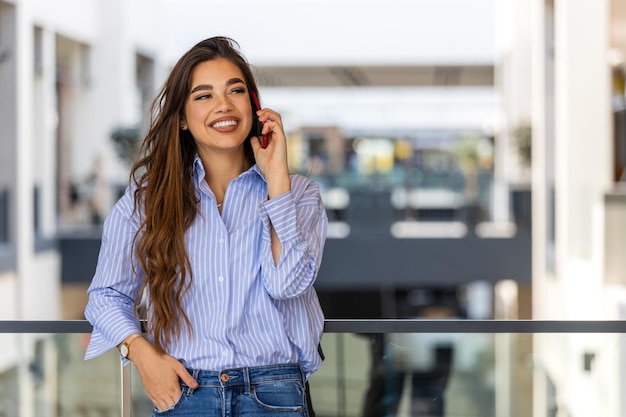 Mulher de negócios jovem feliz e confiante usando telefone celular Mulher profissional sorridente segurando telefone celular de pé no edifício moderno olhando para fora trabalhando fora do escritório