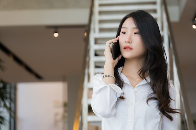 Foto mulher de negócios jovem inteligente, vestindo camisa branca e brim azul, chamando o telefone móvel em pé na frente da escada em conceitual de negócios bem sucedido