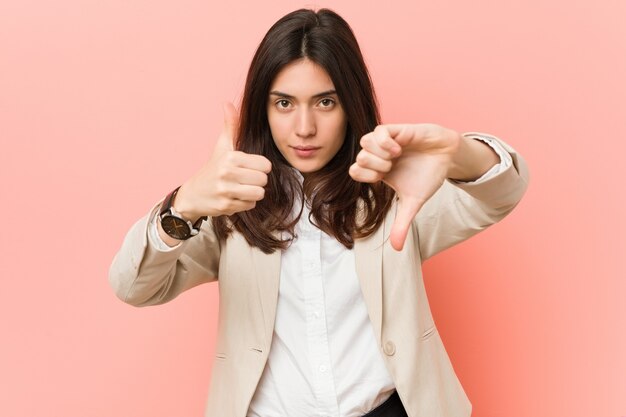 Mulher de negócios jovem morena contra um fundo rosa mostrando os polegares para cima e para baixo, difícil escolher o conceito