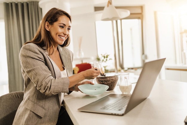 Mulher de negócios jovem multitarefa usando seu laptop e tomando café da manhã enquanto se prepara para ir trabalhar.