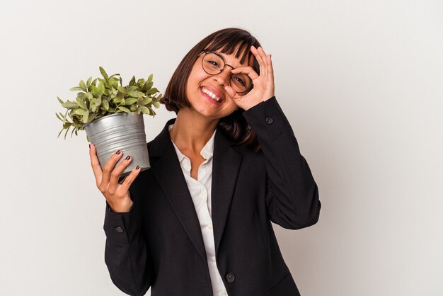 Mulher de negócios jovem raça mista segurando uma planta isolada no fundo branco animado mantendo o gesto ok no olho.