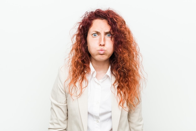 Mulher de negócios jovem ruiva natural isolada contra o fundo branco sopra bochechas, tem expressão cansada