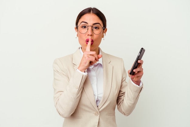 Mulher de negócios jovem segurando um telefone celular isolado no fundo branco, mantendo um segredo ou pedindo silêncio.