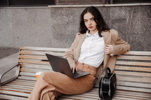 Mulher de negócios jovem sentada em um banco com um laptop do fundo de um prédio de escritórios moderno