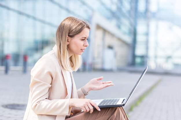 Mulher de negócios jovem sentada na calçada e trabalhando no meio urbano de laptop do centro de escritórios modernos. mulher de negócios, estudante universitária ou faculdade online trabalham ao ar livre. na rua da cidade