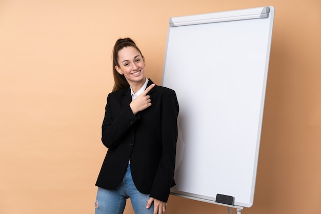 Mulher de negócios jovem sobre parede isolada, dando uma apresentação no quadro branco e apontando para o lado