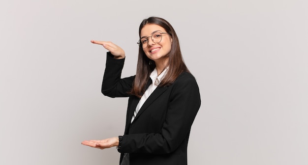 Mulher de negócios jovem sorrindo, sentindo-se feliz, positiva e satisfeita, segurando ou mostrando um objeto ou conceito no espaço da cópia