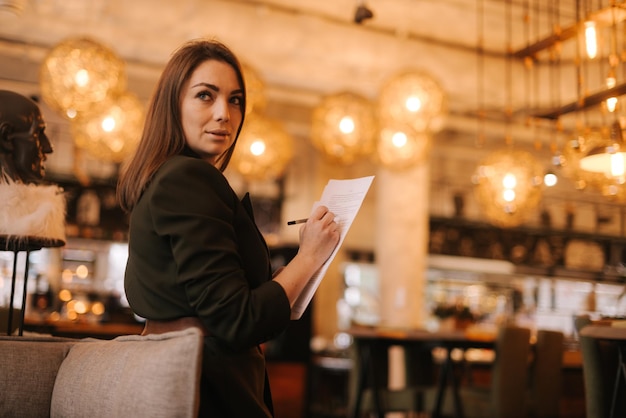 Foto mulher de negócios jovem vestindo roupas formais elegantes no restaurante de luxo moderno segurando documentos em papel e caneta nas mãos senhora de negócios de terno posando no café