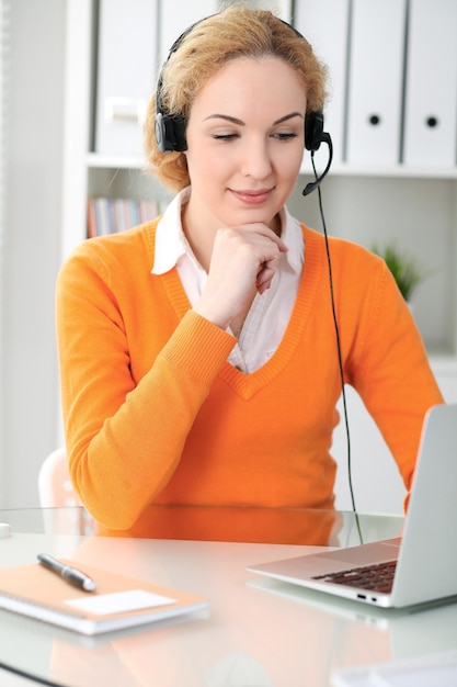 Mulher de negócios linda jovem no fone de ouvido ou operador de chamada se comunica pelo computador portátil. Suéter laranja é uniforme de uma garota.