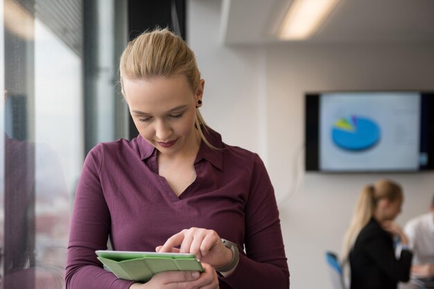 Mulher de negócios loira em roupas casuais, trabalhando no computador tablet no interior do escritório de negócios de inicialização moderna. Grupo de jovens em reunião de equipe desfocado em segundo plano