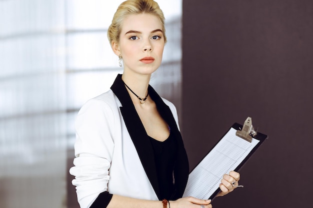 Mulher de negócios loira jovem ou aluna de terno branco está de pé no escritório moderno. Estilo de vida e conceito de pessoas diversas
