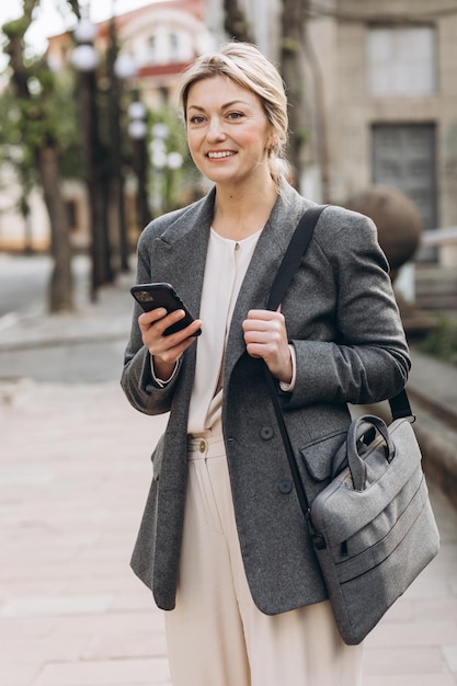 Mulher de negócios loira madura em uma jaqueta cinza sorrindo andando pela cidade e segurando um telefone