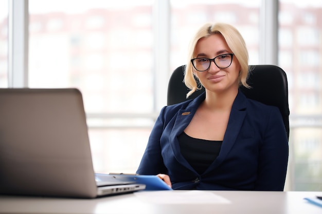 Mulher de negócios loira sorridente com óculos usando laptop no escritório.