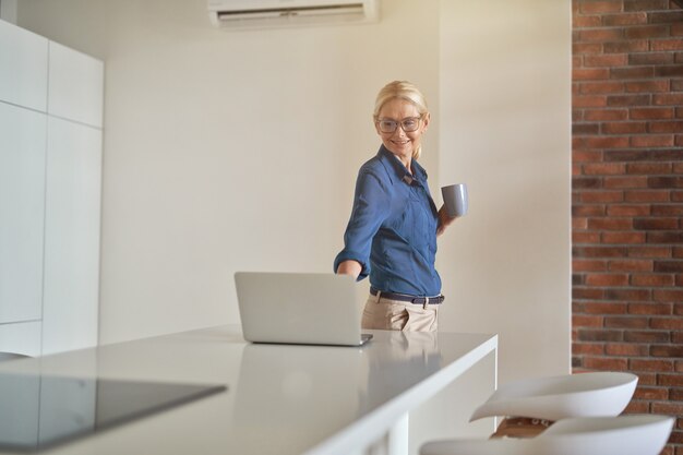 Mulher de negócios madura alegre sorrindo, bebendo café e usando o laptop enquanto está em pé