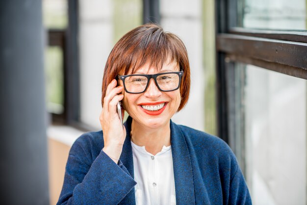 Mulher de negócios mais velha e elegante falando ao telefone em pé dentro de casa perto da janela