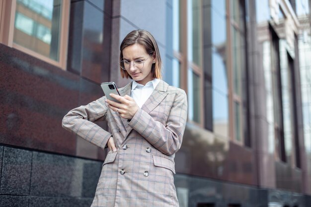 Mulher de negócios moderna jovem em uma jaqueta e óculos, olhando para a tela de um smartphone na cidade