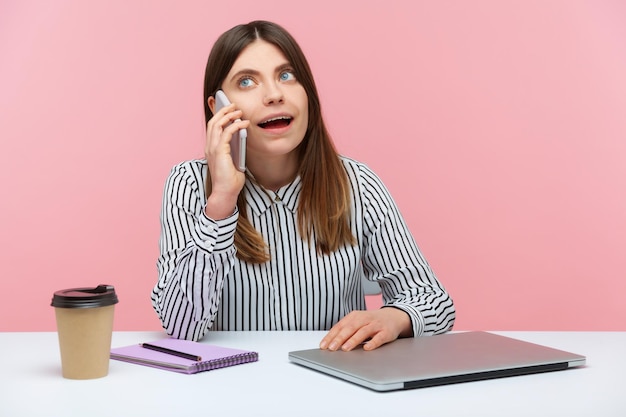 Mulher de negócios morena curiosa em camisa listrada falando telefone sentado no local de trabalho no escritório em casa, tem uma conversa agradável durante o intervalo. Tiro de estúdio interior isolado no fundo rosa