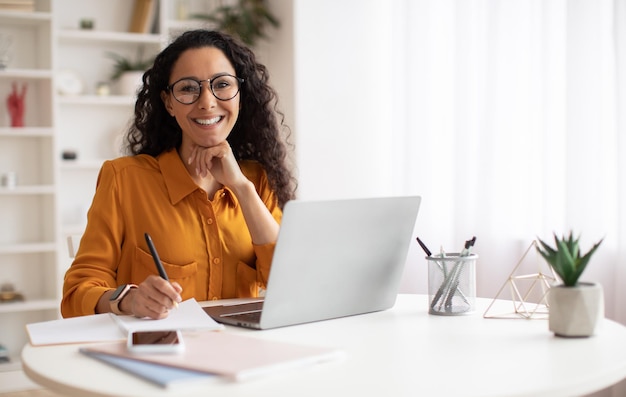 Mulher de negócios morena feliz usando laptop posando sentado no escritório