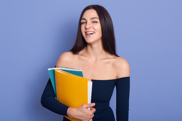 Mulher de negócios morena segurando o papel nas mãos e olhando diretamente para a câmera com um sorriso encantador, usando um vestido elegante