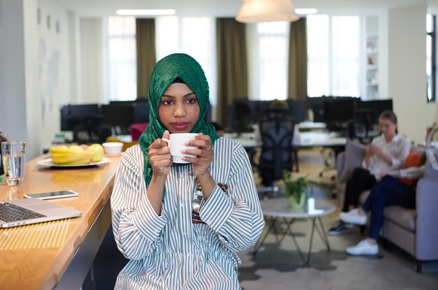 mulher de negócios muçulmana africana vestindo um hijab verde bebendo chá enquanto trabalhava no computador portátil na área de relaxamento no escritório de inicialização de plano aberto moderno. Diversidade, conceito multirracial