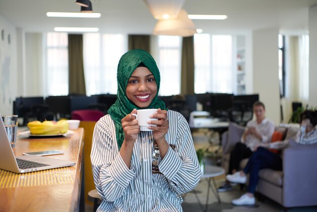 mulher de negócios muçulmana africana vestindo um hijab verde bebendo chá enquanto trabalhava no computador portátil na área de relaxamento no escritório de inicialização de plano aberto moderno. Diversidade, conceito multirracial