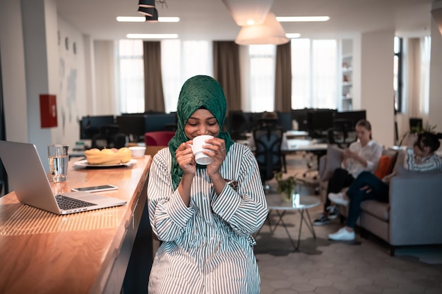 Mulher de negócios muçulmana africana vestindo um hijab verde bebendo chá enquanto trabalhava no computador portátil na área de relaxamento no escritório de inicialização de plano aberto moderno. Foto de alta qualidade