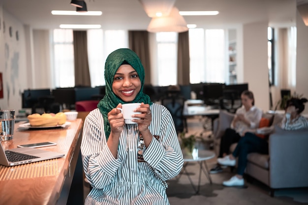 Mulher de negócios muçulmana africana vestindo um hijab verde bebendo chá enquanto trabalhava no computador portátil na área de relaxamento no escritório de inicialização de plano aberto moderno. Foto de alta qualidade